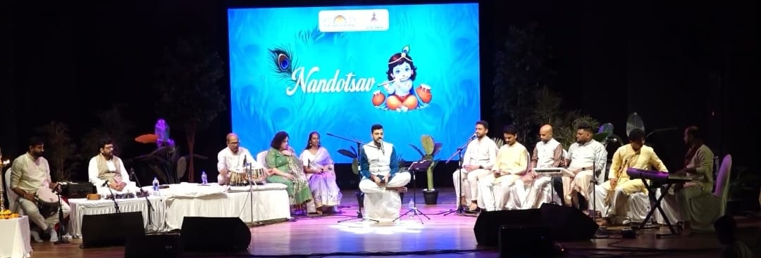 A bhajan ensemble seated on stage with harmonium, drums, and tanpura